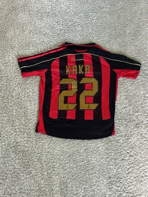 AC Milan matchtröja Kaka #22 - Säljer en klassisk AC Milan matchtröja från Adidas med Kaka och nummer 22 på ryggen. Tröjan är röd och svart med gulddetaljer, Champions League-märke på ärmen och Bwin som sponsor på bröstet. Perfekt för fotbollsfans som vill sticka ut på läktaren eller planen.