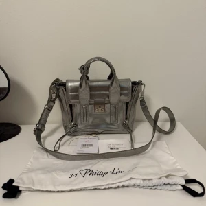 3.1 Phillip Lim Mini Satchel - Silverfärgad handväska från 3.1 Phillip Lim med både handtag och avtagbar axelrem.   Köpte från Vestiaire Collective 2 år sen men har inte använt så mycket, därför säljer den.  Hade betalt extra för Authentication & Quality Control (se bilden) så det är äkthetbevis för väskan.   Säljer för 1000kr och kom gärna med bud.  Kan hämta i Tyresö.