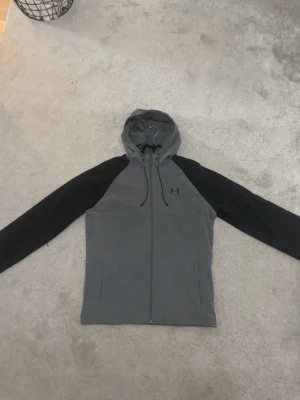 Grå och svart vindjacka Under Armour - Extremt skön och bekväm vindjacka från Under Armour i grått med svarta ärmar. Jackan har dragkedja framtill, två fickor och justerbar huva med snören. Använd ungefär 1-2 gånger så extremt bra skick. Tillverkad i lätt och slitstarkt material.