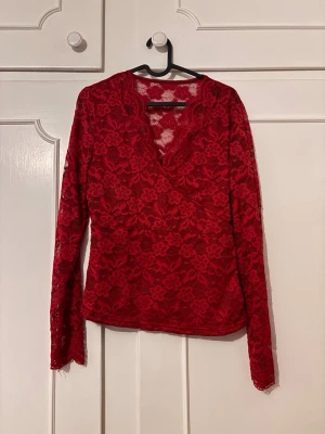 Röd spetsblus med v-ringning - Snygg röd blus i spets med blommigt mönster och v-ringad omlottfront. Långärmad modell som är lätt transparent och har fina vågiga kanter vid halsen. 