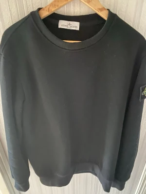 Stone Island tjock tröja  - Svart stickad tröja från Stone Island med rund hals och ribbade muddar. Tröjan har den klassiska logotypen på vänster ärm och är tillverkad i ett mjukt material som känns skönt mot huden. Enkel och stilren design som passar till många olika looks.