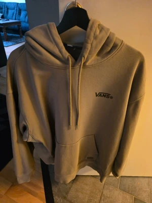 Beige hoodie från Vans med logga - Snygg beige hoodie från Vans med svart logga på bröstet. Hoodien har en stor huva med dragsko och en klassisk magficka. Tillverkad i mjukt material som känns skönt mot huden. Perfekt för dig som gillar streetwear och enkel stil.