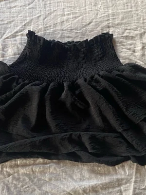 Svart volangkjol med smock - Supersöt svart kortkjol med smock i midjan och volangig nederdel. Kjolen har en luftig känsla och passar perfekt till en trendig vardagslook. Enkel att matcha med olika toppar och sneakers.