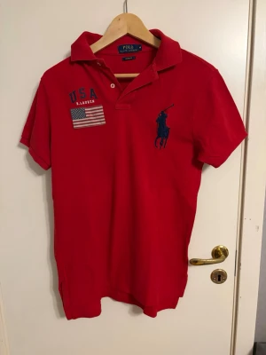Röd pikétröja från Polo Ralph Lauren - Snygg röd pikétröja från Polo Ralph Lauren med blå logga, USA-tryck och amerikansk flagga på bröstet. Klassisk krage och korta ärmar. Custom fit-modell som passar perfekt till en avslappnad stil.