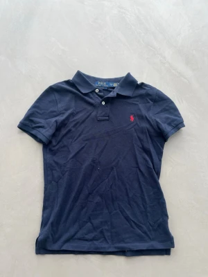 Marinblå Ralph Lauren pikétröja från Zalando - OBS! KOMMER ATT STRYKAS INNAN FRAKTAT OCH DEFEKTEN SOM SYNS PÅ BILD KOMMER GÅ BORT EFTER RENSNING. PIKÉT ÄR ÄKTA! Klassisk marinblå pikétröja från Ralph Lauren med korta ärmar och krage. Tröjan har två knappar framtill och den ikoniska röda logotypen broderad på bröstet. Tillverkad i bomull för en bekväm och stilren look. Passar dem runt 165-175. 