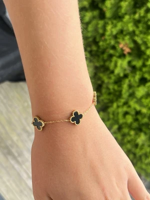 Guldarmband med svarta klöverhängen - Snyggt armband i guldton med tre svarta klöverformade hängen, omgivna av små guldfärgade pärldetaljer. Kedjan är tunn och elegant, vilket ger en stilren och trendig look. Perfekt accessoar för dig som gillar klassiska men ändå moderna smycken.
