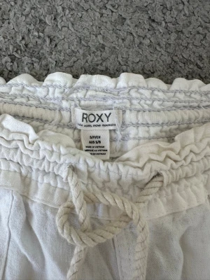 Vita linnebyxor från Roxy - Säljer ett par vita linnebyxor från Roxy med resår och dragsko i midjan. Byxorna har en bakficka med broderad logga och är tillverkade i ett luftigt linnematerial. Perfekta för en avslappnad och stilren look. Dom har en lagning på insida lår kan skriv så skickar jag bild på det! 