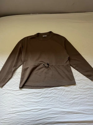 Brun sweatshirt med dragsko i midjan - Mjuk brun sweatshirt från Cubus med rund halsringning och lång ärm. Tröjan har en snygg dragsko i midjan som ger en justerbar passform. Perfekt för en avslappnad och trendig look.