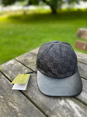 Svart Gg keps med GG-mönster - Säljer en svart keps från Gg med det klassiska GG-monogrammet över hela kepsen. Skärmen är i svart läder och resten av kepsen är i tyg. Snygg och stilren accessoar som lyfter vilken outfit som helst.