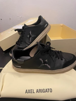Svarta sneakers Axel Arigato - Snygga svarta sneakers från Axel Arigato med broderad fågel på sidan och texten 1.40MM. Skorna har vita sulor och är tillverkade i läder med svarta skosnören. Perfekta för en trendig streetwear-look.