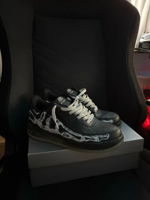 Nike Air Force 1 skelleton - Säljer ett par svarta Nike Air Force 1 sneakers med vita flames längs sidorna och vita skosnören. Klassisk låg modell med perforerad tåbox och Nike-logga på hälen. Skorna har en robust gummisula och är tillverkade i läder.