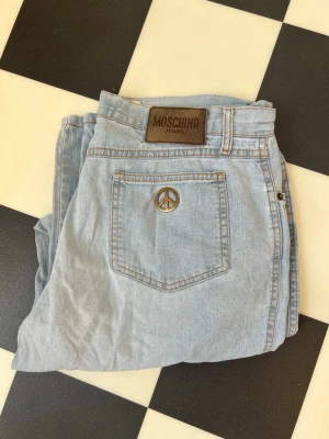 Ljusblå Moschino jeans med peace-märke - Säljer ett par ljusblå jeans från Moschino med klassisk femficksmodell och ett coolt peace-märke på bakfickan. Jeansen har en brun läderpatch med Moschino-logga i midjan och är gjorda i mjuk denim. Perfekta för dig som gillar vintage och streetstyle.