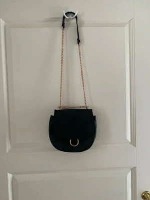 Svart axelväska med guldkedja - Snygg svart axelväska i rundad form med guldfärgad kedjerem och dekorativ ringdetalj på locket. Väskan är tillverkad i slätt konstläder och har en stilren, minimalistisk design som passar till många outfits. Kolla gärna mina andra annonser❗️