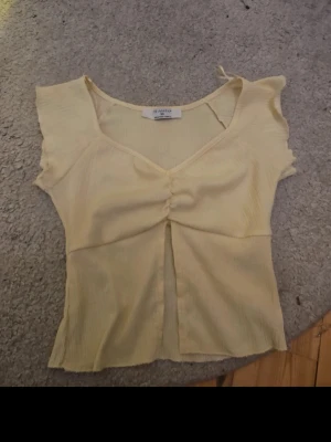Ljusgul ribbad topp från Primark - Söt ljusgul topp från Primark i ribbad bomullsmix. Toppen har korta ärmar, v-ringning och en snygg rynkdetalj framtill. Perfekt för dig som gillar en feminin och enkel stil.