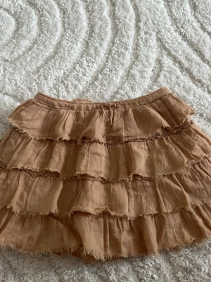 Beige volangkjol från Zara - Superfin beige kortkjol från Zara med flera lager volanger och råa kanter. Kjolen har en mjuk resår i midjan och är tillverkad i ett lätt och luftigt material som passar perfekt till varmare dagar. Men en inbyggt byxa