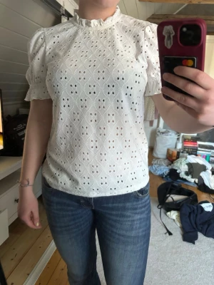 Vit broderad blus med puffärm - Söt vit blus med broderie anglaise-mönster och puffiga korta ärmar. Blusen har liten volangkrage och smockdetaljer vid ärmslut. Tillverkad i bomullstyg som känns lätt och luftig. Perfekt för en romantisk och trendig look.