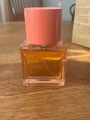 CAIA Senso parfym med rosa kork - En snygg parfymflaska från CAIA med doften Senso. Flaskan är fyrkantig i genomskinligt glas med en varm orange vätska och har en stor, matt rosa kork. Modern och stilren design som passar perfekt på sminkbordet.
