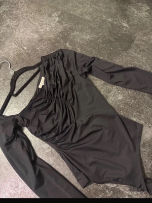 Svart långärmad body med rynk - Snygg svart body med långa ärmar och rynkdetaljer framtill. Tillverkad i ett stretchigt, mjukt material som sitter skönt mot kroppen. Perfekt för att skapa en stilren och trendig look. Knäppning i grenen för enkel på- och avtagning.