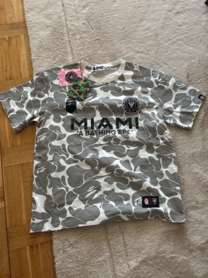 Grå camo Inter Miami t-shirt BAPE - Säljer en grå och vit camouflagemönstrad t-shirt från A Bathing Ape i samarbete med Inter Miami CF. T-shirten har tryck fram med MIAMI och klubbmärke, samt klassiska BAPE-detaljer. Skön passform och perfekt för dig som gillar streetwear och fotboll.