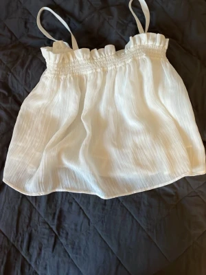 Vit linnetopp från MNG med volang - Söt vit linnetopp från MNG med tunna axelband och volangdetalj upptill. Toppen har en luftig och lätt struktur, perfekt för varma dagar. Smockad upptill för en bekväm passform och en flowy känsla.