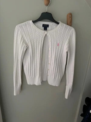 Vit kabelstickad kofta Polo Ralph Lauren - Kabelstickad vit kofta från Polo Ralph Lauren med rund halsringning och knappar framtill. Klassisk design med rosa broderad logga på bröstet. Långa ärmar och ribbade muddar ger en snygg och tidlös look.