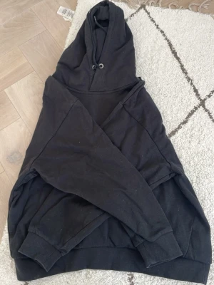 Svart hoodie med huva - En klassisk svart hoodie med huva och ribbade muddar vid ärmslut och nederkant. Hoodien har dragsko i huvan och är tillverkad i mjukt material, perfekt för en avslappnad och cool look. Passar dig som gillar enkel och stilren design.