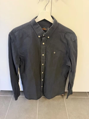 Svart skjorta från Morris stl L 41/42 - Säljer en stilren svart skjorta från Morris med klassisk button-down krage och vita knappar. Skjortan har ett diskret broderat emblem på bröstet och är tillverkad i mjuk bomull. Perfekt för dig som gillar en clean och tidlös look.