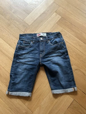 Levi's 511 slim blå jeansshorts  - Säljer ett par klassiska blå jeansshorts från Levi's med fem fickor, uppvikta benslut och normal passform. Shortsen har gylf med knapp och dragkedja samt Levi's ikoniska läderpatch bak i midjan. Perfekta för sommaren!