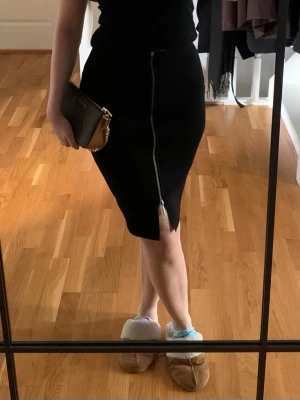 Svart pennkjol med dragkedja - Säljer en svart pennkjol med en snygg silverfärgad dragkedja framtill som går hela vägen ner och avslutas med en liten slits. Kjolen har en figurnära passform och är knälång. Perfekt att styla med en enkel topp och boots. Passar även S, stretchigt material.