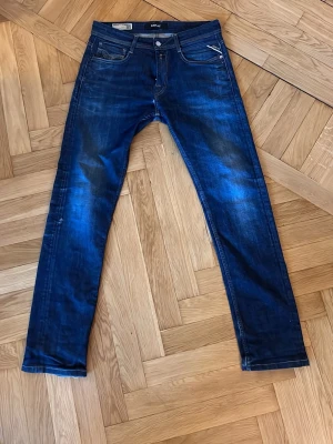Mörkblå Replay Roccos - Snygga mörkblå jeans från Replay med raka ben och klassisk femficksdesign. Jeansen har coola slitningar på framsidan och detaljerade bakfickor, jeansen är i storlek 30/34 men är upplagda till 30/32. Bland de mer ”loose” sittande jeansen från Replay