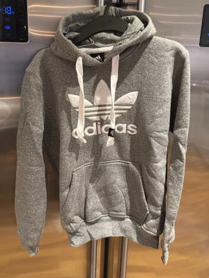 Grå hoodie från Adidas Originals - Säljer en grå hoodie från Adidas Originals med stor vit logga och klassiska trebladet på bröstet. Hoodien har huva med vita snören, ribbade muddar och en stor magficka. Perfekt för dig som gillar sportig och avslappnad stil.