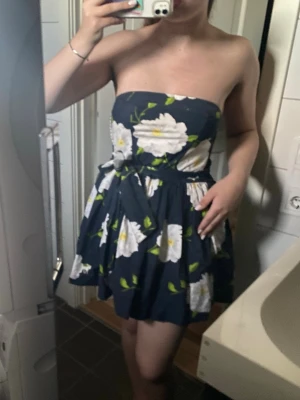 Blommig axelbandslös kortklänning - Axelbandslös kortklänning med mörkblå bas och stora vita blommor med gröna detaljer. Klänningen har en knyt i midjan och en utsvängd kjol. Perfekt för sommarens varma dagar och ger en fräsch och ungdomlig look.