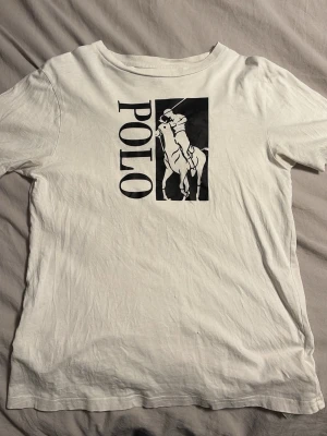 Vit t-shirt från Polo Ralph Lauren - Vit t-shirt med svart POLO-tryck och ikonisk hästlogga på bröstet. Klassisk passform och rund halsringning. Perfekt för en stilren och avslappnad look.