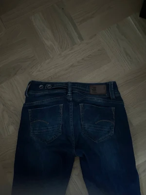 Mörkblå bootcut jeans G-Star - Snygga mörkblå jeans från G-Star med bootcut passform och klassiska bakfickor med broderade detaljer och coola knappar. Jeansen har dubbla knappar i midjan och är tillverkade i ett slitstarkt denimtyg. Perfekta för en trendig och avslappnad stil.