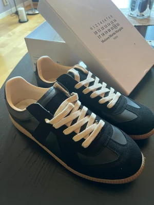 Maison Margiela Gats svarta  - Maison Margiela gats sneakers i svart läder och mocka med vita skosnören och beige sula. Klassisk low-top siluett med retrovibbar och diskret branding på plösen. Perfekta för dig som gillar stilrena och tidlösa skor.