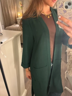 Mörkgrön tygkappa lång kofta - Stilren mörkgrön kappa från Forever 21 med raka ärmar och två stora framfickor. Kappan har en enkel, öppen design utan knappar och är tillverkad i ett mjukt tyg som ger en skön känsla. 