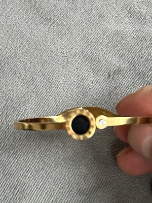 18K guldpläterat Guldigt armband med svart sten - Snyggt armband i guldfärgad metall med en rund svart sten i mitten och en liten klar detalj bredvid. Stilren och modern design som passar perfekt till din outfit. Armbandet har ett smidigt lås och känns lätt på handleden.