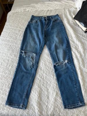 Blå ripped jeans från Gina Tricot - Säljer ett par blå mom fit jeans från Gina Tricot med slitningar och hål på båda benen. Klassisk femficksmodell med dragkedja och knapp. Perfekta för en avslappnad och trendig look.
