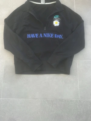 Svart Nike tröja med tryck - Svart tröja från Nike med halv dragkedja, blå text 'Have a Nike Day' på bröstet och en vit blomma. Stor turkos Nike-logga på ryggen och ett litet gult peace-märke. Tillverkad i mjukt material och har lång ärm. Perfekt för dig som gillar streetwear och snygga detaljer.