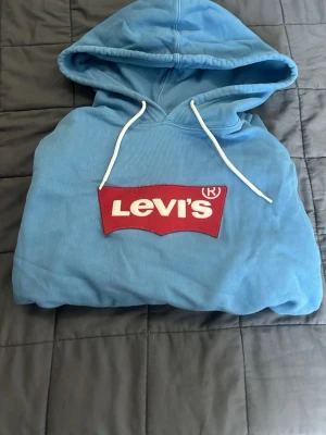 Blå hoodie från Levi's med logga - Snygg blå hoodie från Levi's med stor röd logga på bröstet och vita snören i huvan. Klassisk passform med känguruficka framtill och ribbade muddar. Perfekt för dig som gillar streetwear och vill ha något bekvämt och stilrent. Har tappat lite färg men det märks knappast.     Pris kan diskuteras