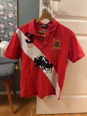 Röd pikétröja från Polo Ralph Lauren - Röd pikétröja med vit diagonal rand och broderade detaljer från Polo Ralph Lauren.        Det är barnstorlek XL men sitter precis som M för herr.             Nypris ca 1600, unik. Bra skick 