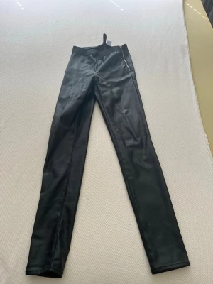 Svarta leggings i skinnimitation - Säljer ett par svarta leggings i skinnimitation med hög midja och smal passform. De har en dragkedja på sidan och är tillverkade i ett glansigt material som ger en cool look. Perfekta för dig som vill ha ett edgy och trendigt plagg.