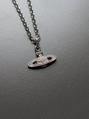 Vivienne Westwood halsband med orb - Snyggt halsband från Vivienne Westwood med den ikoniska orb-berlocken i silverfärg. Berlocken är dekorerad med små gnistrande stenar och hänger på en kraftig kedja. Perfekt för dig som gillar statement-accessoarer och cool design.