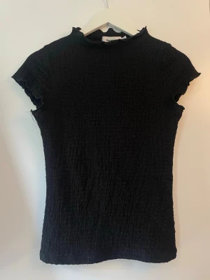 Svart ribbad topp från Lager 157 - Säljer en svart ribbad topp från Lager 157 med korta ärmar och lätt rynkade kanter vid hals och ärmslut. Toppen har en figurnära passform och är tillverkad i ett stretchigt material som ger en bekväm känsla. Perfekt att styla med jeans eller kjol. 