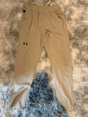 Beige mjukisbyxor från Under Armour - Snygga beige mjukisbyxor från Under Armour med logotyp på vänster ben. Byxorna har en relaxed fit, elastisk midja med dragsko och sidofickor. Perfekta för chill eller träning, tillverkade i mjukt och stretchigt material. De har en liten liten brännskada som man kan se på andra bilden.