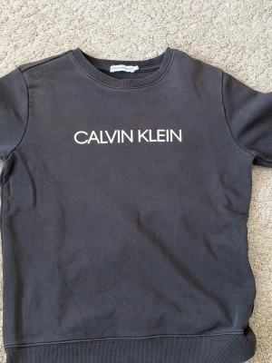 Svart sweatshirt från Calvin Klein - Cool svart sweatshirt från Calvin Klein med vit logga framtill och rund hals. Tröjan har långa ärmar och ribbade muddar vid ärmslut och nederkant. Perfekt till jeans  för en avslappnad look.