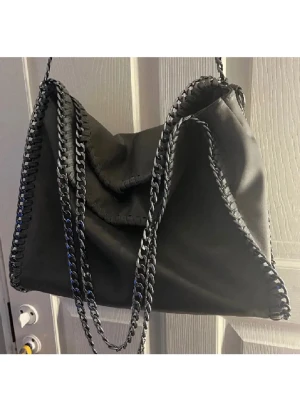 Svart axelväska med kedjedetaljer - Cool svart axelväska med kedjedetaljer längs kanterna och axelremmen. Väskan har en mjuk och slouchy form, vilket ger en avslappnad vibe. Materialet ser ut att vara syntetiskt och väskan har en modern design som sticker ut.