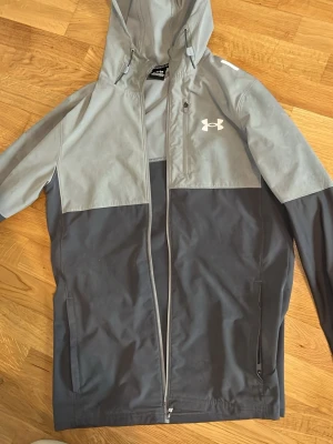 Grå vindjacka från Under Armour - Snygg tvåfärgad vindjacka från Under Armour i ljusgrått och mörkgrått. Jackan har huva med dragsko, hel dragkedja och två sidofickor med dragkedja. Materialet är lätt och vattenavvisande, perfekt för blåsiga dagar. Logotypen syns på bröstet.