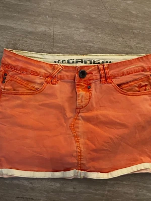 Orange kortkjol - Säljer en cool orange kortkjol från gang med vita detaljer längs kanterna. Kjolen har klassiska jeansfickor fram och bak, knapp och dragkedja. Perfekt för dig som vill ha en färgstark och unik look. Kjolen är mer korall färgad än det ser ut på bilderna. Midjemåttet är 41cm