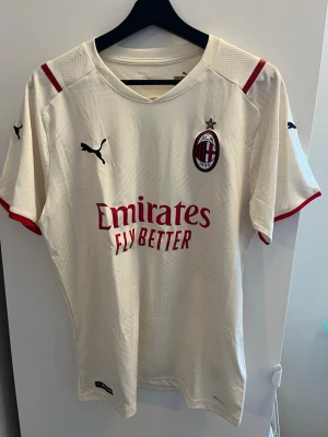 AC Milan bortatröja och shorts Puma - Säljer en AC Milan bortatröja och shorts från Puma i ljusbeige med röda detaljer. Tröjan har klubbens emblem på bröstet, Puma-loggor på ärmar och bröst samt sponsortryck 'Emirates Fly Better' på framsidan. Materialet är lätt och andas, perfekt för fotboll eller träning. Tröja M Shirts XL
